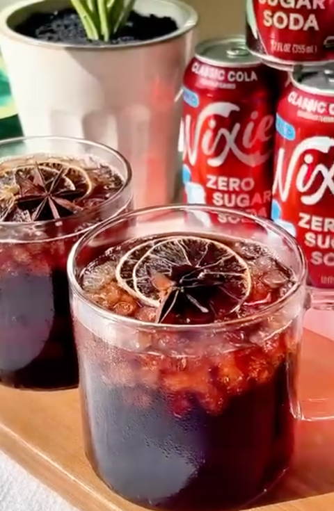 Two glasses of Chai-Cherry Cola Cooler garnished with dried citrus and star anise, with Nixie Classic Cola cans in the background.