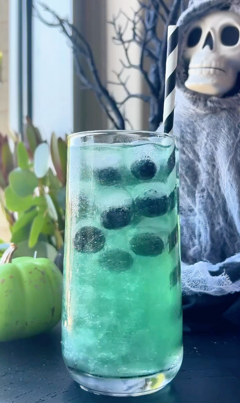 Green Halloween mocktail with blueberries and a striped straw, surrounded by spooky décor including a skeleton and green pumpkin.
