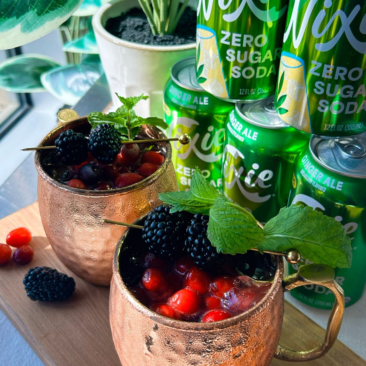 Two copper mugs filled with iced berry drinks, blackberries, cranberries and fresh mint, with cans of Nixie Ginger Ale Organic Zero Sugar Soda in the background.
